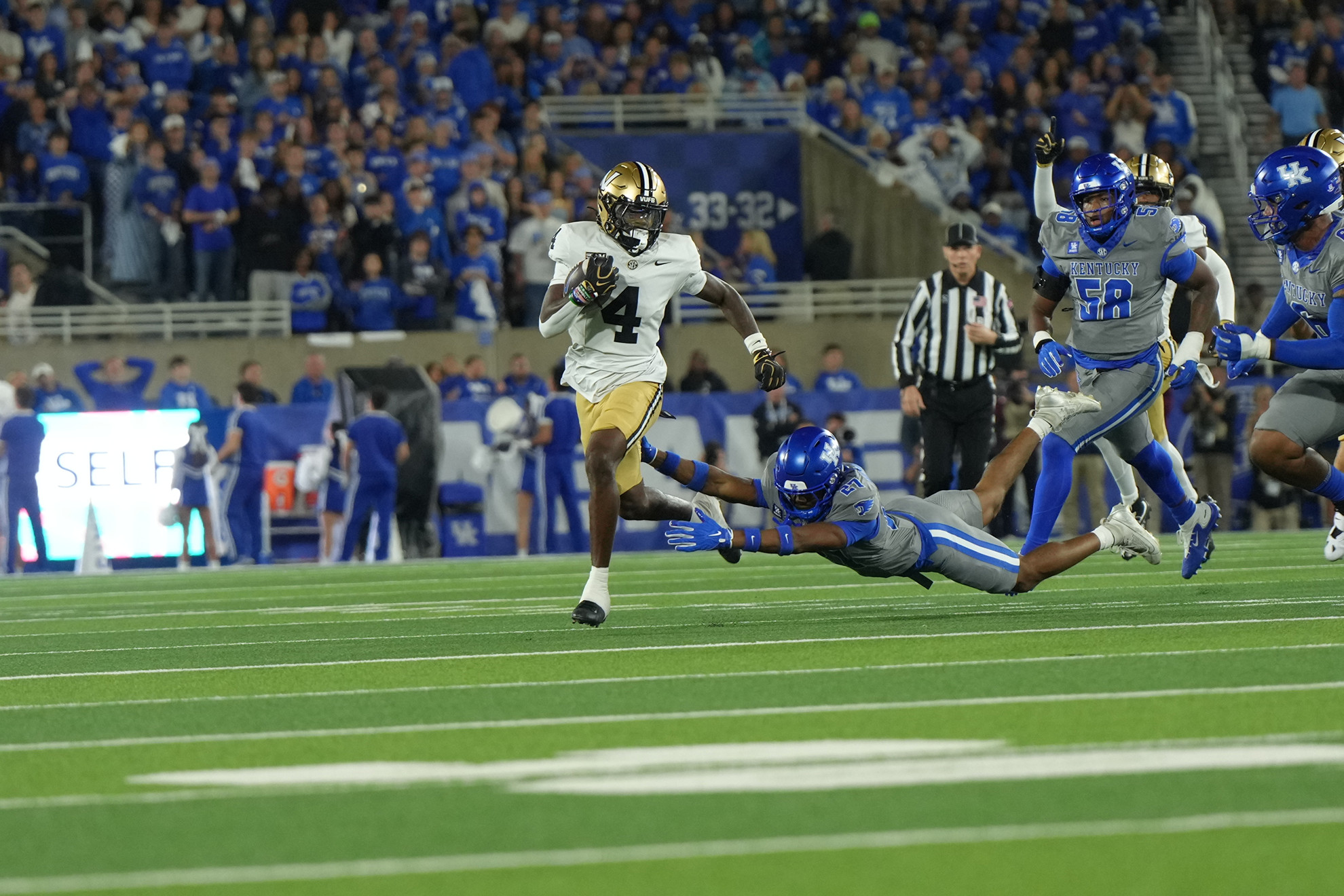 A football player in a white jersey with the number 4 runs down the field as a defender in a blue uniform dives to tackle him.