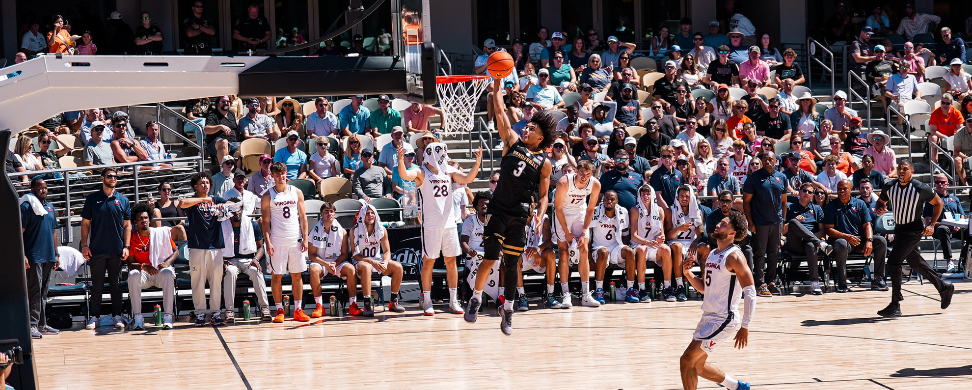 A basketball player in a black jersey leaps to shoot the ball while opponents and a crowd watch from the stands and sidelines.