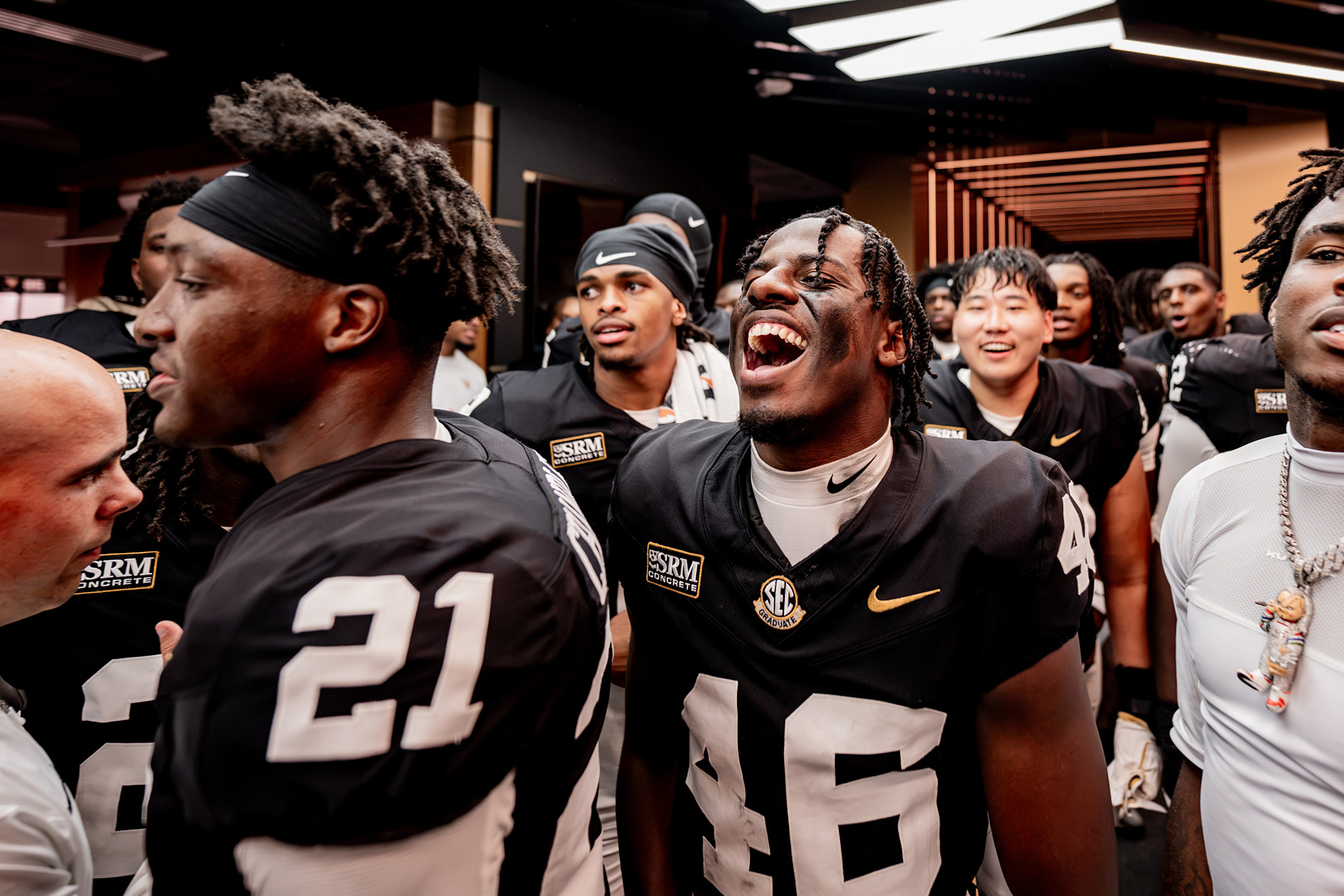 A group of football players in black uniforms with white numbers celebrate together in a locker room, with one player laughing loudly.