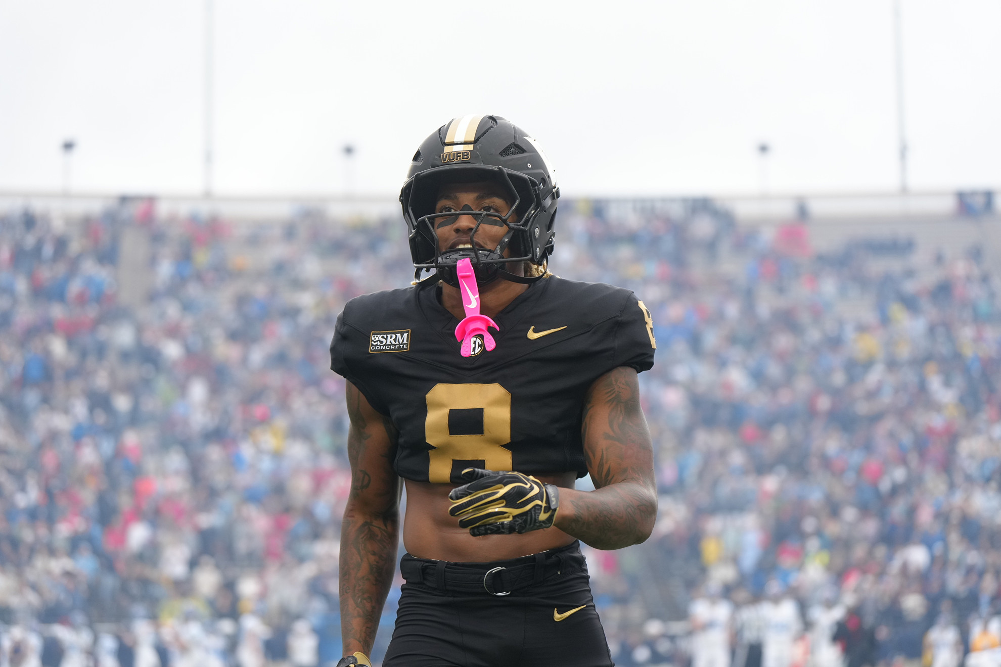 An athlete in a black and gold uniform with the number 8 stands on a football field, wearing a helmet and gloves, with a pink object hanging from their neck, as a large crowd fills the stadium behind them.