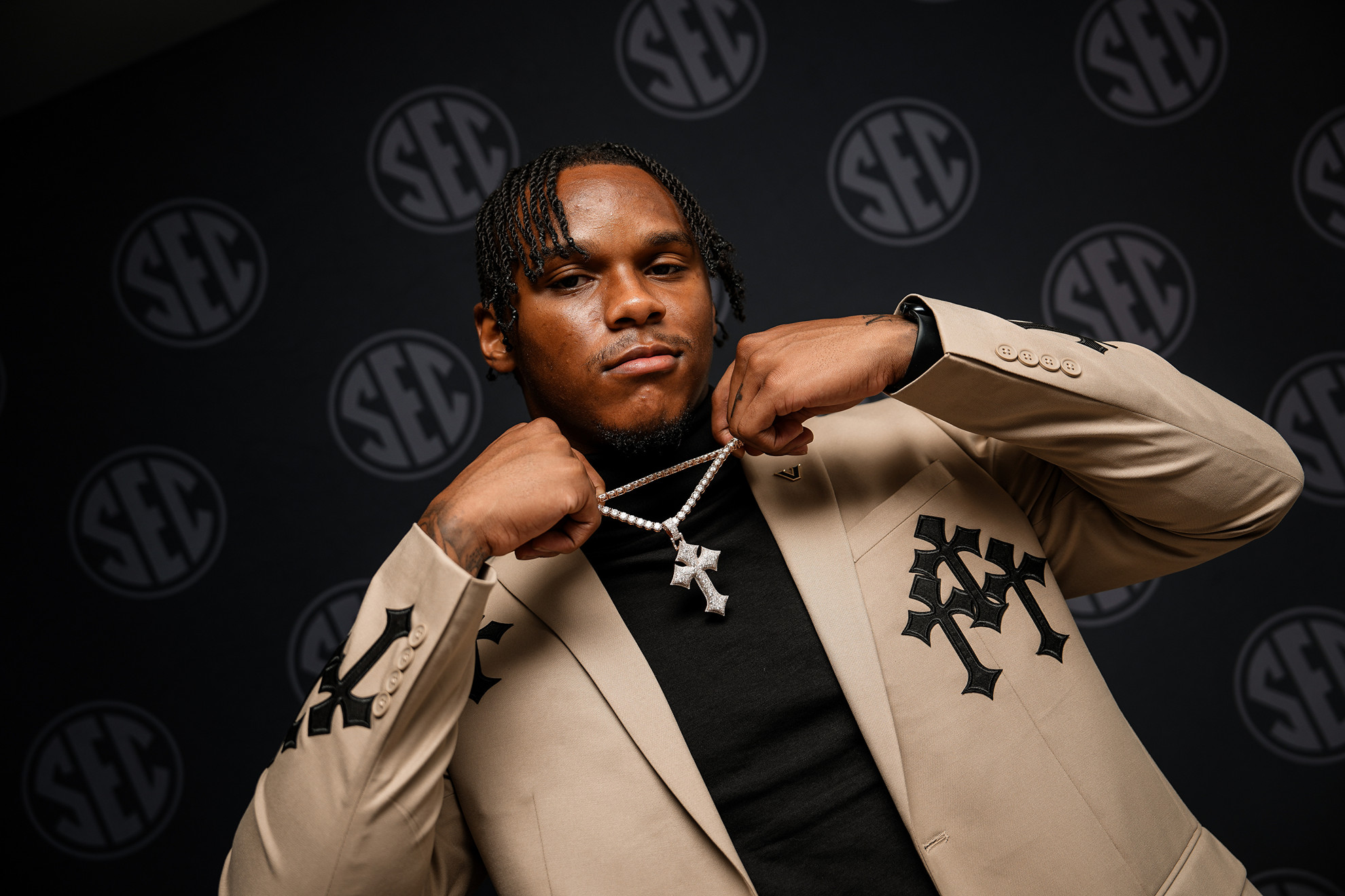 A person in a beige blazer with black embroidery adjusts a diamond-encrusted cross necklace against a backdrop featuring repeated SEC logos.