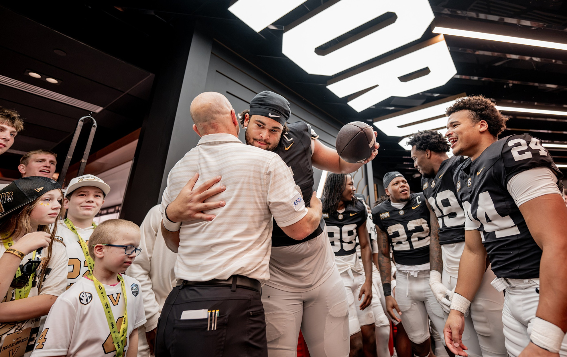 A football player in a black jersey holds a football while embracing a man in a white shirt, surrounded by teammates and young fans in a brightly lit indoor space.