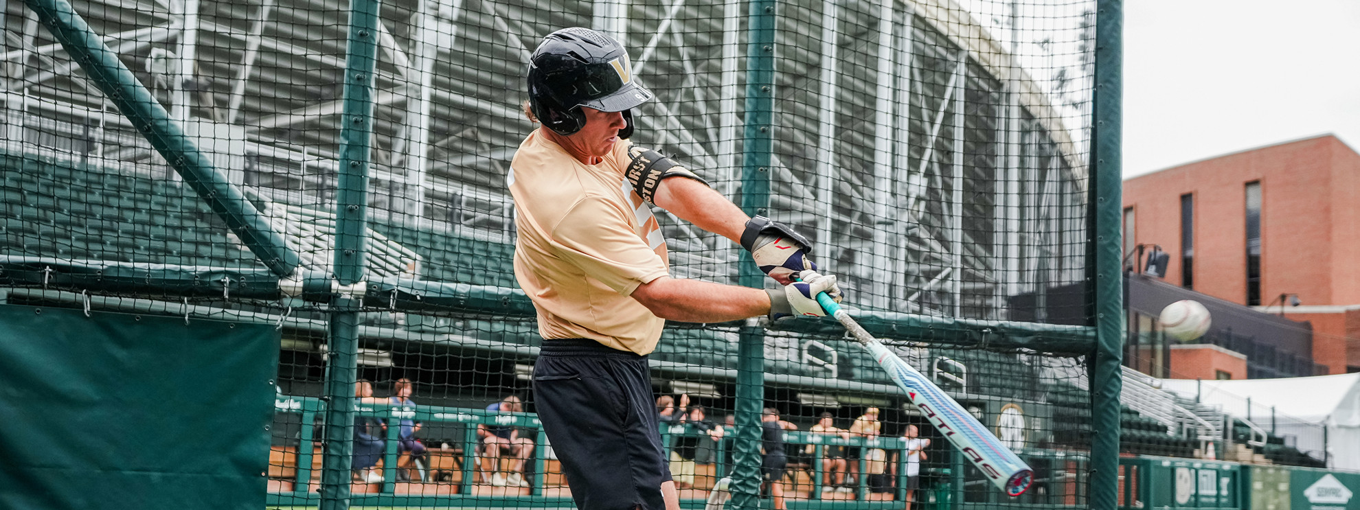 A person wearing a helmet and batting gloves swings a baseball bat in a batting cage, with a stadium visible in the background.