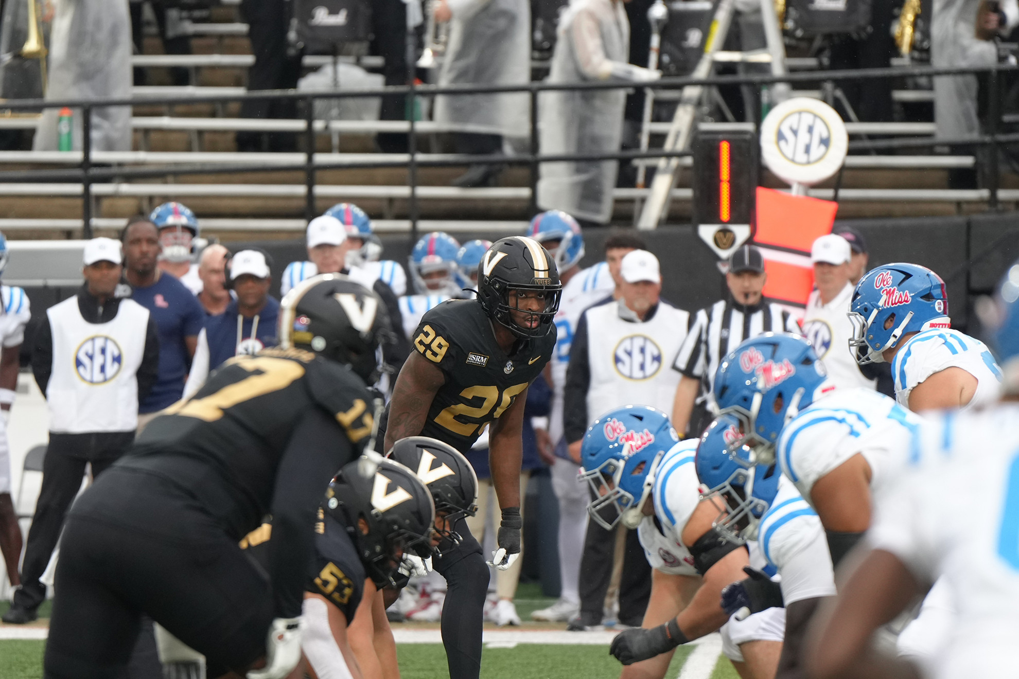 A football player in a black and gold uniform with the number 29 stands at the line of scrimmage, ready for play, while opponents in blue and white gear are positioned across from him.