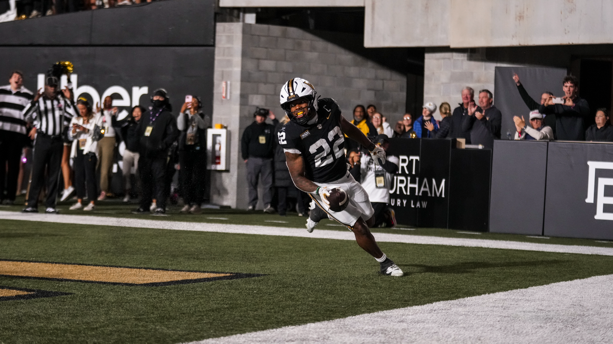 An athlete in a black and white uniform runs with a football across the field during a nighttime game, with spectators and staff visible in the background.