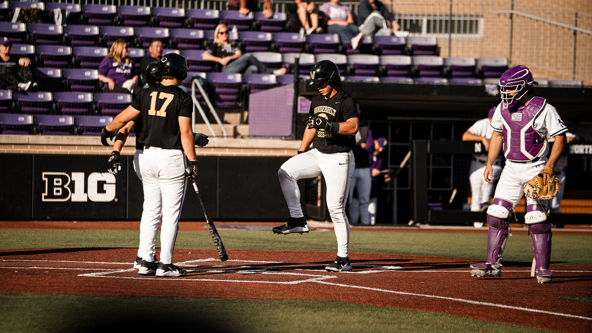 A baseball player in a black uniform with the number 17 stands at home plate, while another player in a black uniform and a catcher in purple gear stand nearby on the field during a game.