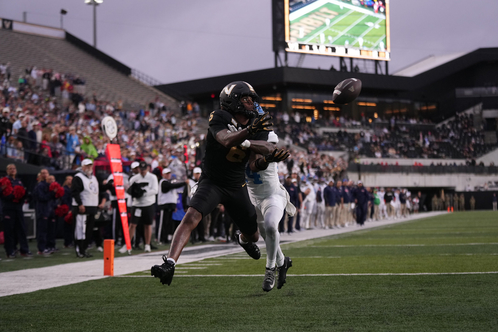 A football player in black dives to catch the ball as a defender in white attempts to tackle him during a game.