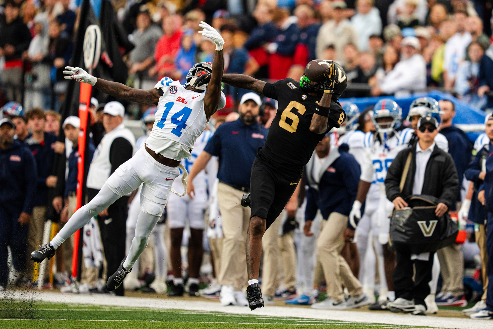 Two football players leap in mid-air, one in white jersey number 14 and another in black jersey number 6, during a game with spectators in the background.